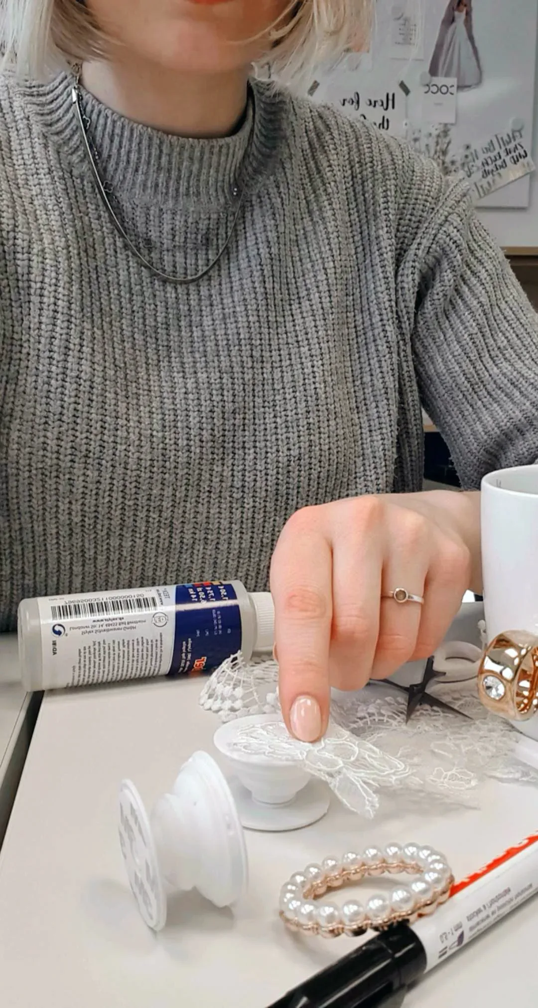 Eine Frau mit hellblonden Haaren und grauem Strickpullover bereitet ein DIY-Projekt vor. Sie deutet auf ein weißes Popsocket, das mit Spitze verziert wird, umgeben von Kleber, Perlen und Bastelmaterialien.