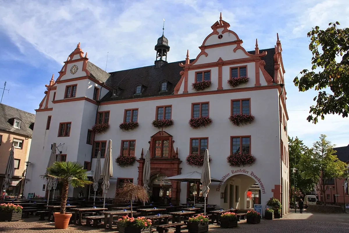 Das historische Alte Rathaus in Darmstadt, eine prächtige weiße Fassade mit roten Akzenten und einem kleinen Uhrturm. Vor dem Gebäude stehen Tische und Stühle auf einem gepflasterten Platz.