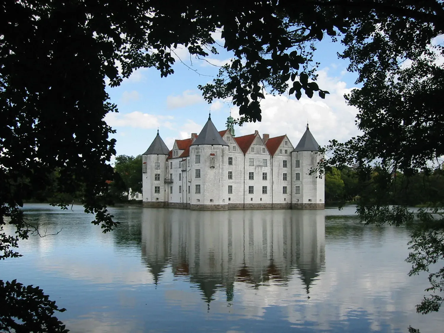 Das weiße Wasserschloss Glücksburg, umgeben von einem ruhigen See, spiegelt sich im Wasser. Umrahmt von grünen Bäumen, hebt sich das Schloss vor dem blauen Himmel ab.
