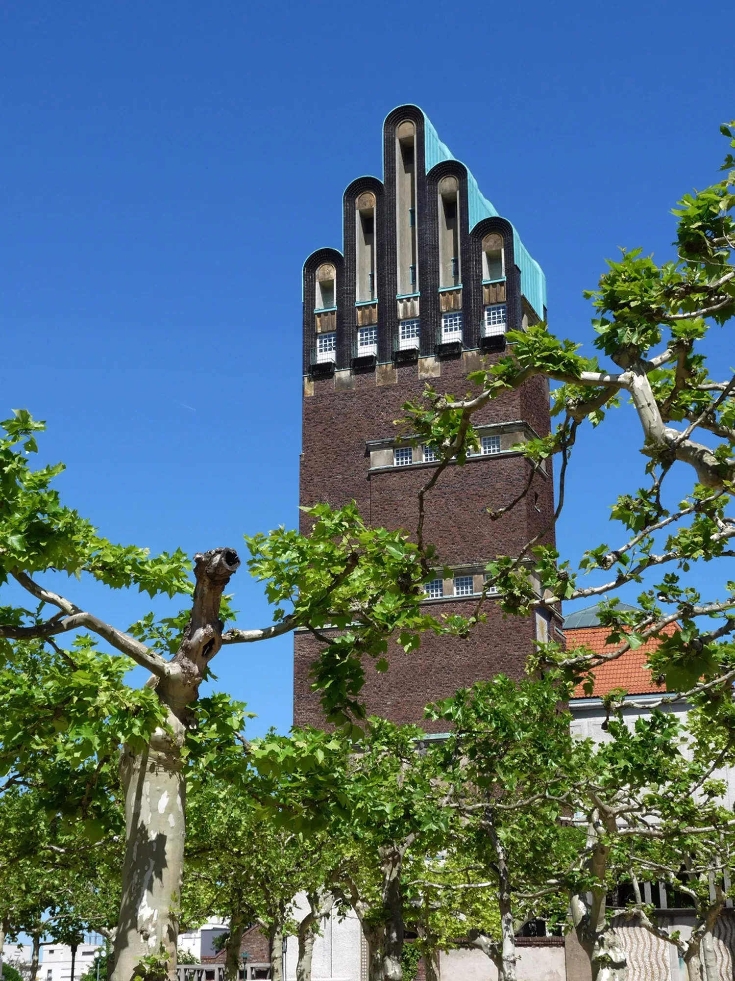Der Hochzeitsturm in Darmstadt mit seinem markanten Stufenturm-Design, umgeben von grünen Bäumen und einem strahlend blauen Himmel im Hintergrund.