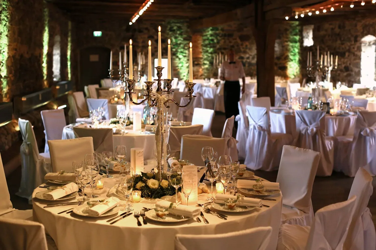 Eleganter Hochzeitstisch mit weißen Stuhlhussen und Kerzenleuchtern in rustikalem Ambiente.