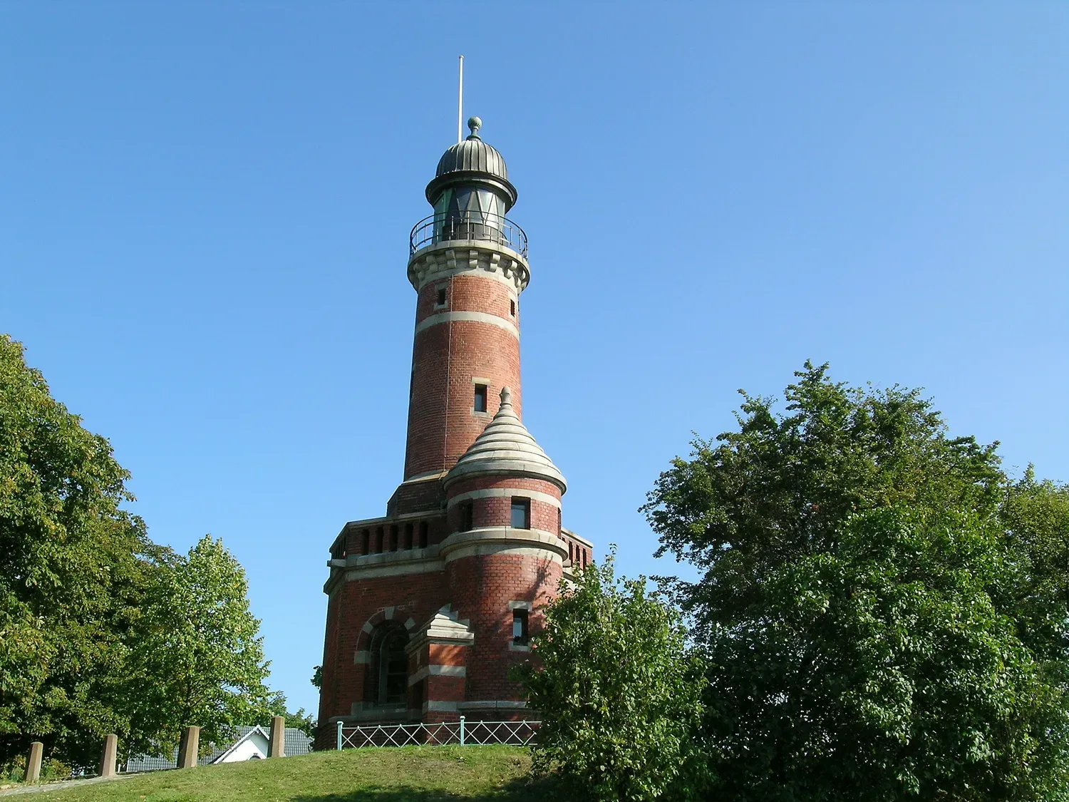 Ein historischer Backstein-Leuchtturm in Kiel-Holtenau, der unter einem strahlend blauen Himmel steht, umgeben von grünen Bäumen.