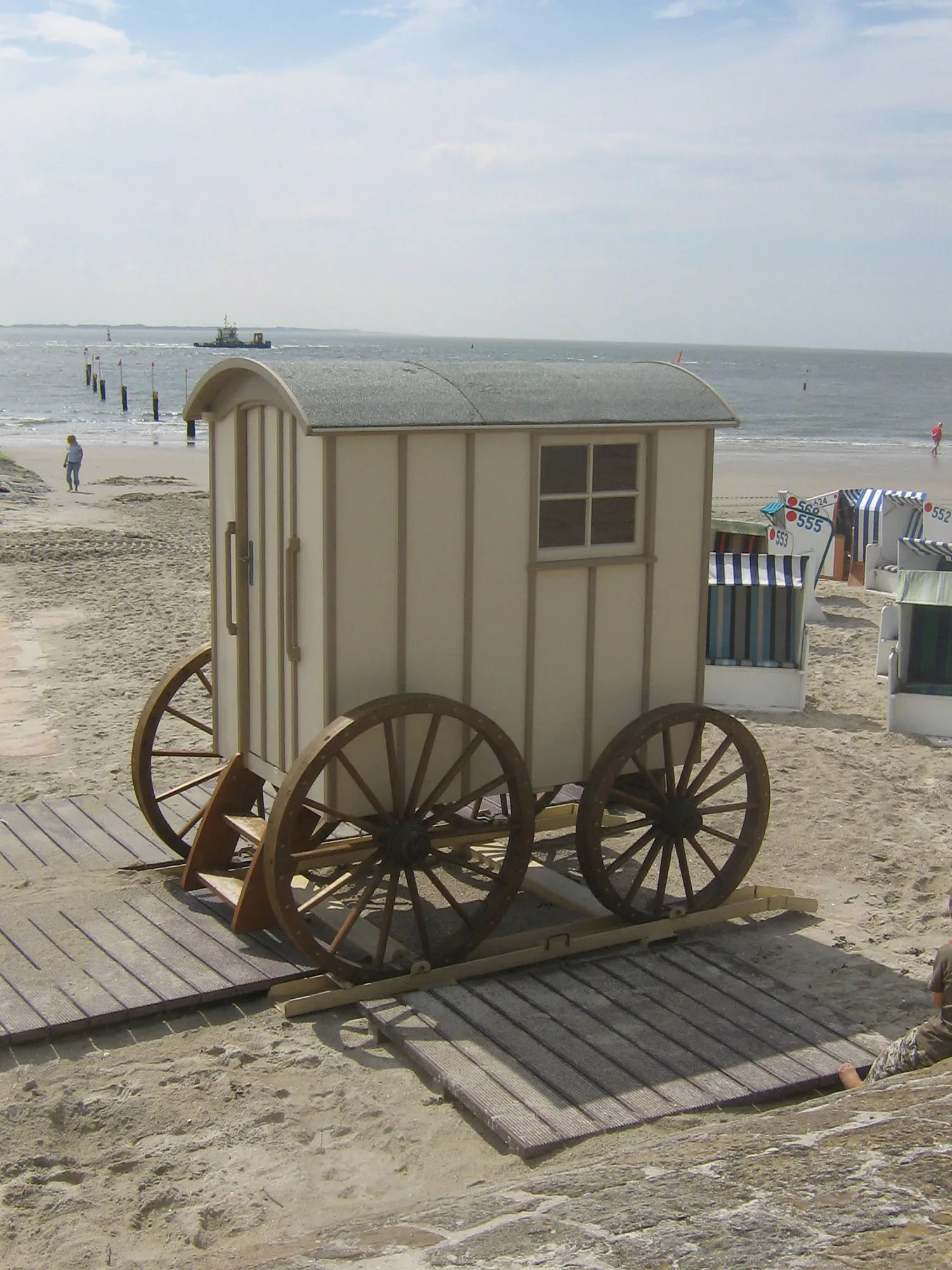 Eine historische Strandkabine auf Rädern, die 'Hochtiedsstuv', steht am Sandstrand von Norderney. Im Hintergrund sind die Nordsee und weitere Strandkörbe zu sehen.