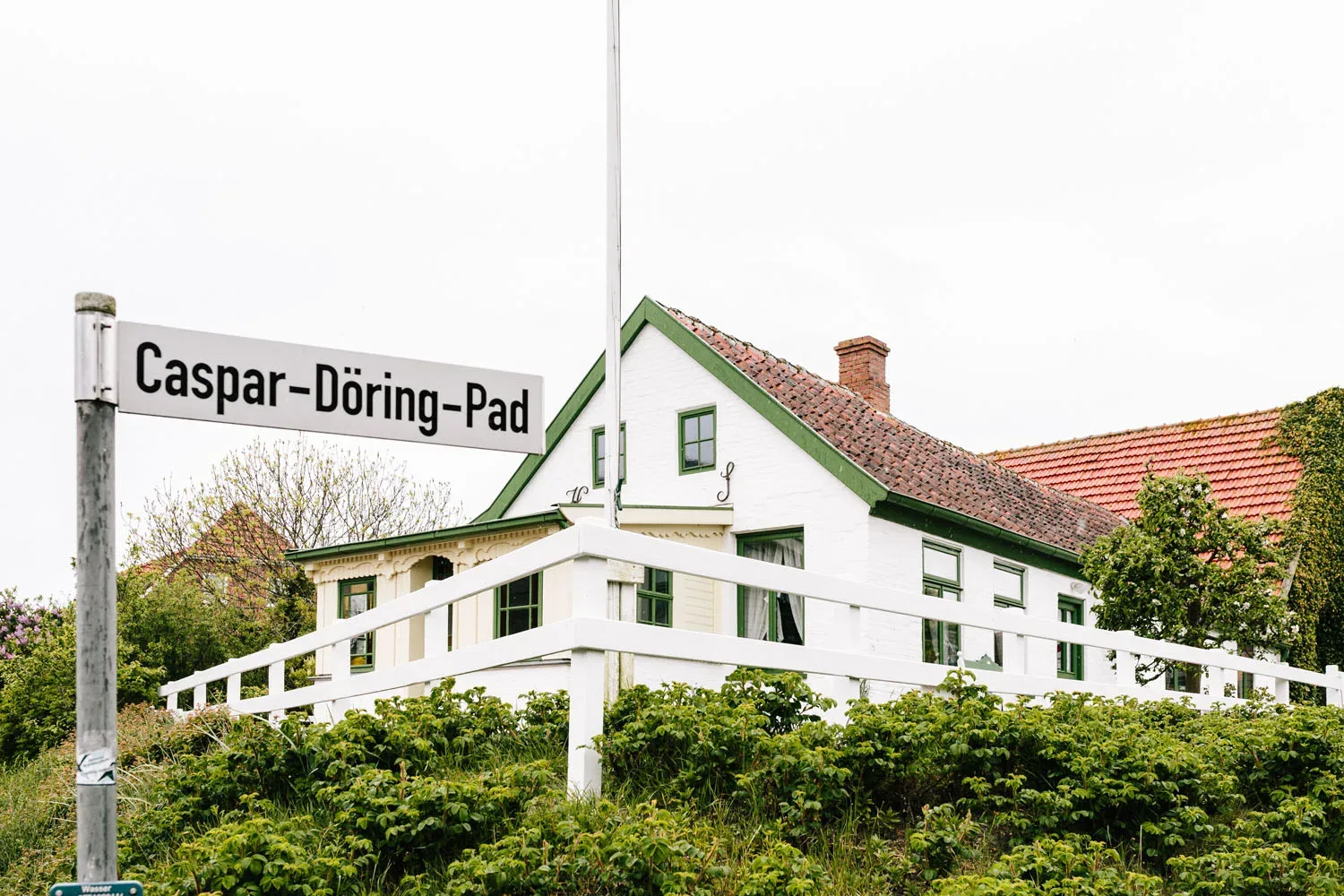 Weiß gestrichenes Haus mit grünen Fensterrahmen und Ziegeldach am Caspar-Döring-Pad, umgeben von grüner Vegetation und einem weißen Zaun. Ein Straßenschild mit der Aufschrift "Caspar-Döring-Pad" ist im Vordergrund zu sehen.