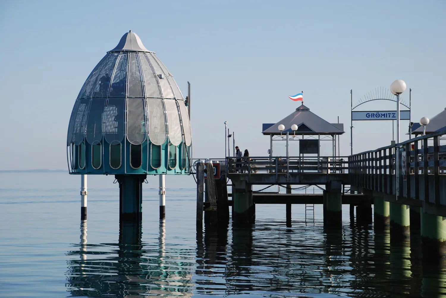 Blick auf die Tauchgondel an der Seebrücke in Grömitz, eine futuristische Konstruktion mit gläserner Kuppel, die über dem ruhigen Wasser der Ostsee schwebt, mit einem Steg im Hintergrund.