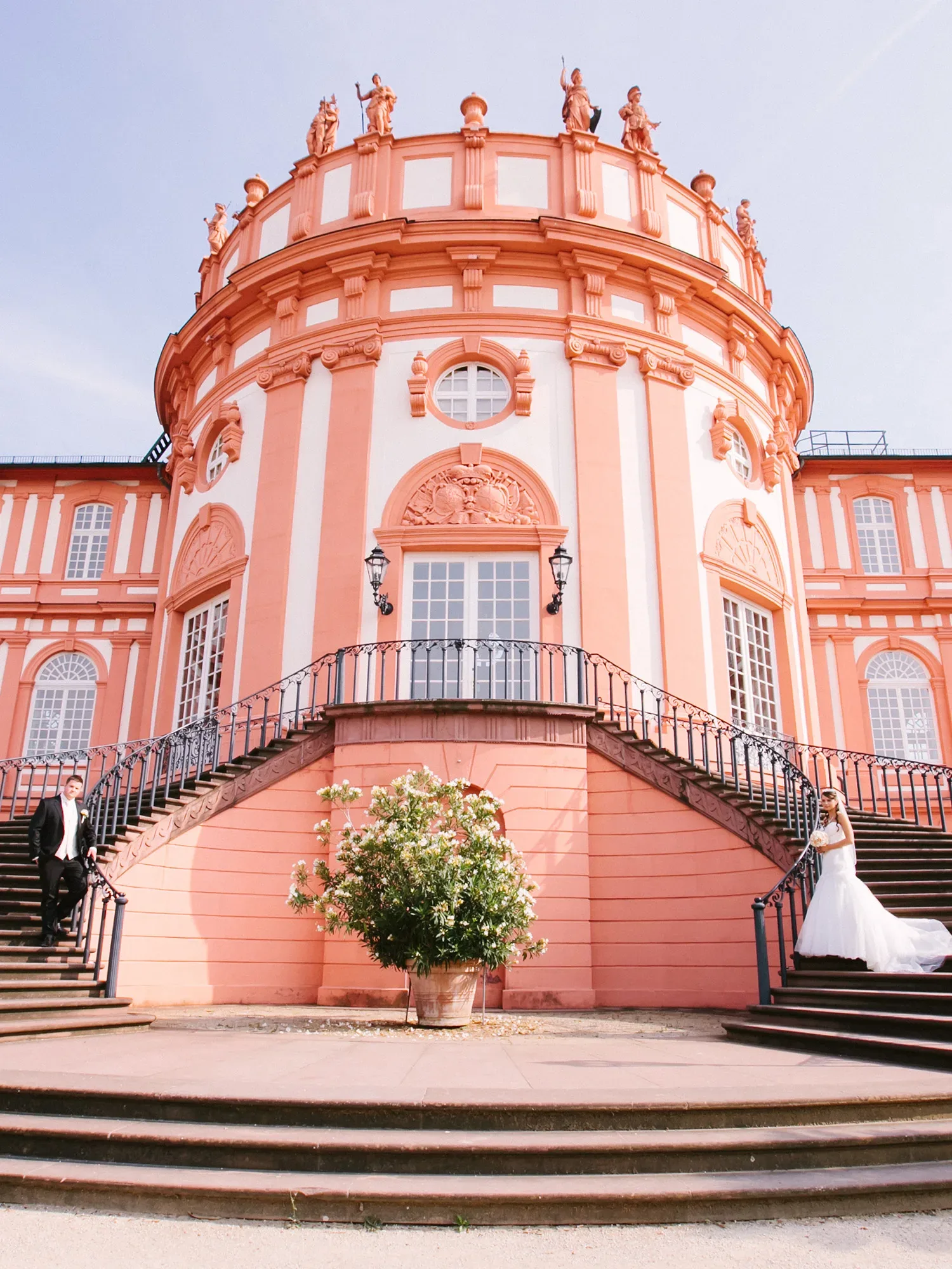 Die großartige Fassade des rosafarbenen Schloss Biebrich in Wiesbaden dient als Kulisse für ein Brautpaar auf den Treppen, umgeben von kunstvollen Details und strahlendem Sonnenschein.