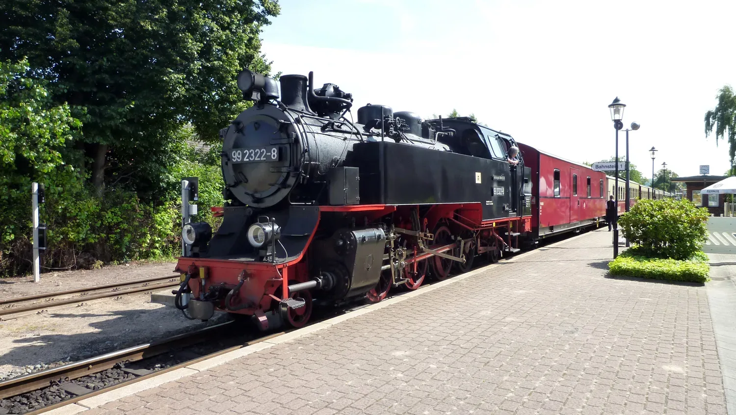 Eine historische Dampflokomotive mit der Nummer 99 2322-8 steht am Bahnsteig, bereit zur Abfahrt, während ein Schaffner aus dem Fenster schaut. Die Lokomotive ist schwarz-rot lackiert und zieht rote Waggons hinter sich her.