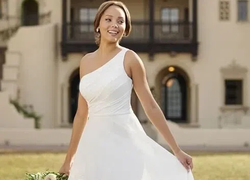 Braut in einem einseitig schulterfreien, eleganten A-Linien-Kleid, lächelt und posiert vor einer historischen Villa. Der fließende Rock schwingt sanft.