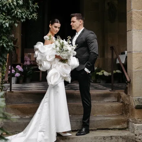 Elegante Braut im weißen Kleid mit voluminösen Ärmeln und üppigem Blumenstrauß, posiert neben Bräutigam in dunklem Anzug vor historischer Kulisse.