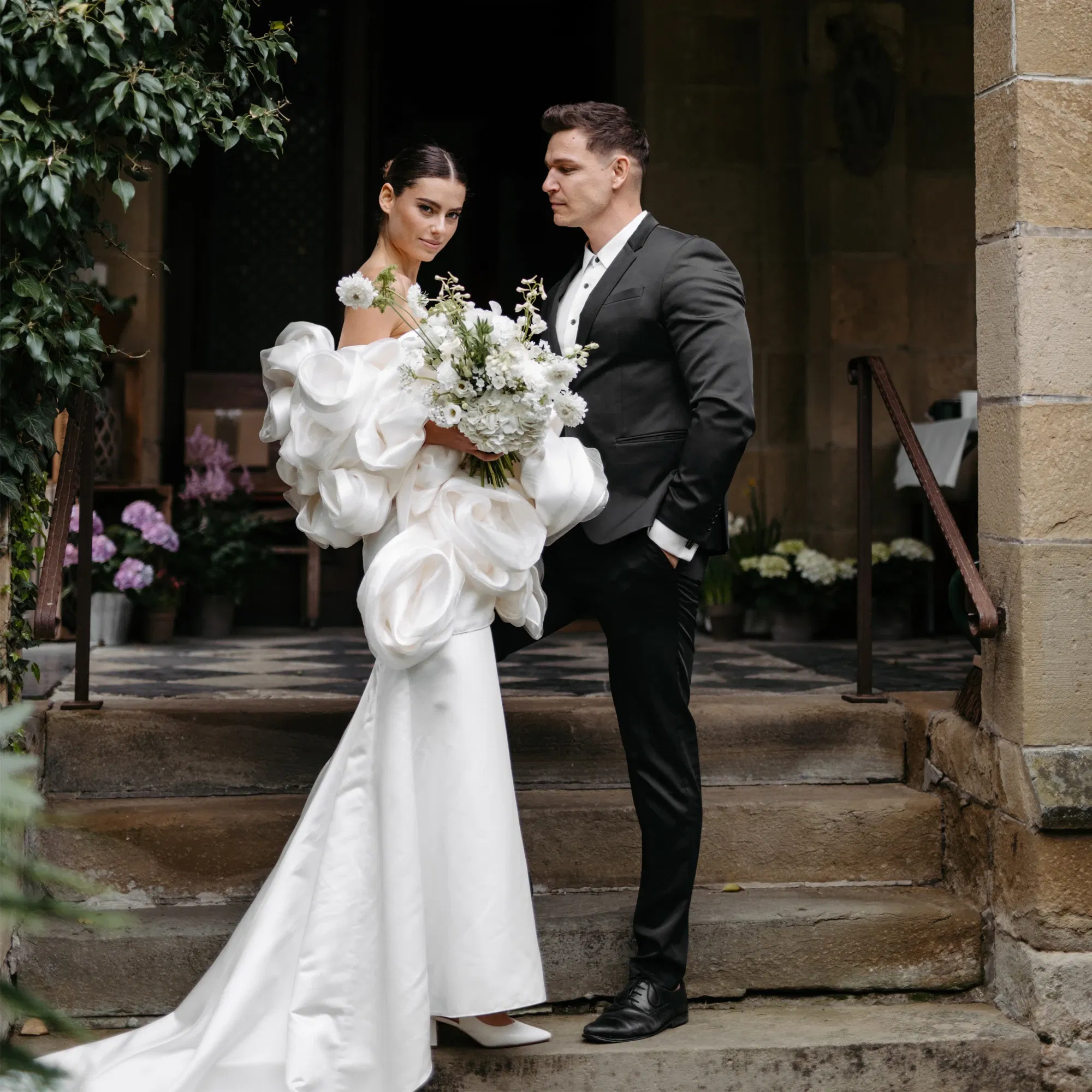 Elegante Braut im weißen Kleid mit voluminösen Ärmeln und üppigem Blumenstrauß, posiert neben Bräutigam in dunklem Anzug vor historischer Kulisse.