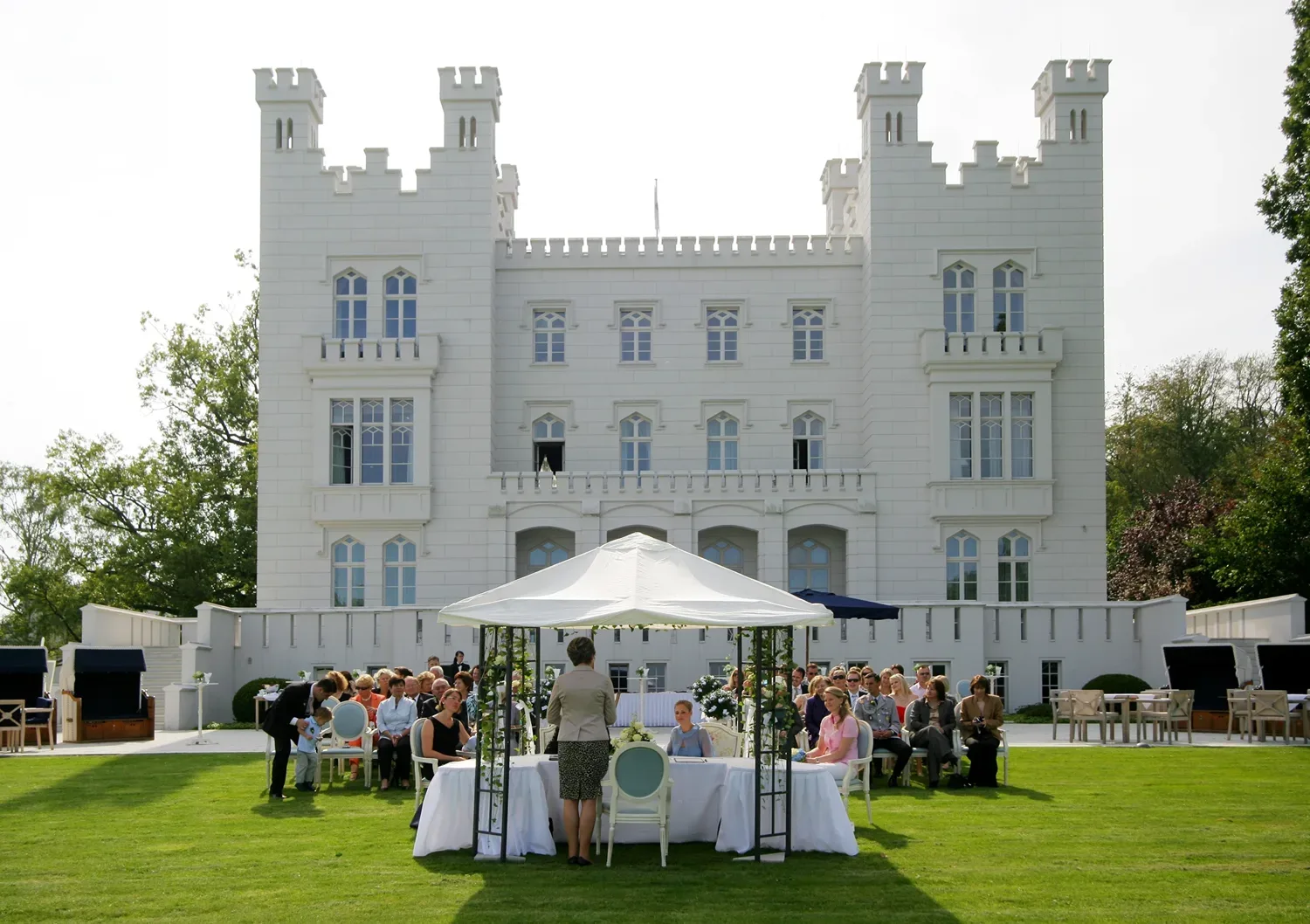 Eine Hochzeitszeremonie im Freien vor einem großen weißen Schloss. Gäste sitzen unter einem dekorativen Pavillon, während die Zeremonie im Garten stattfindet.