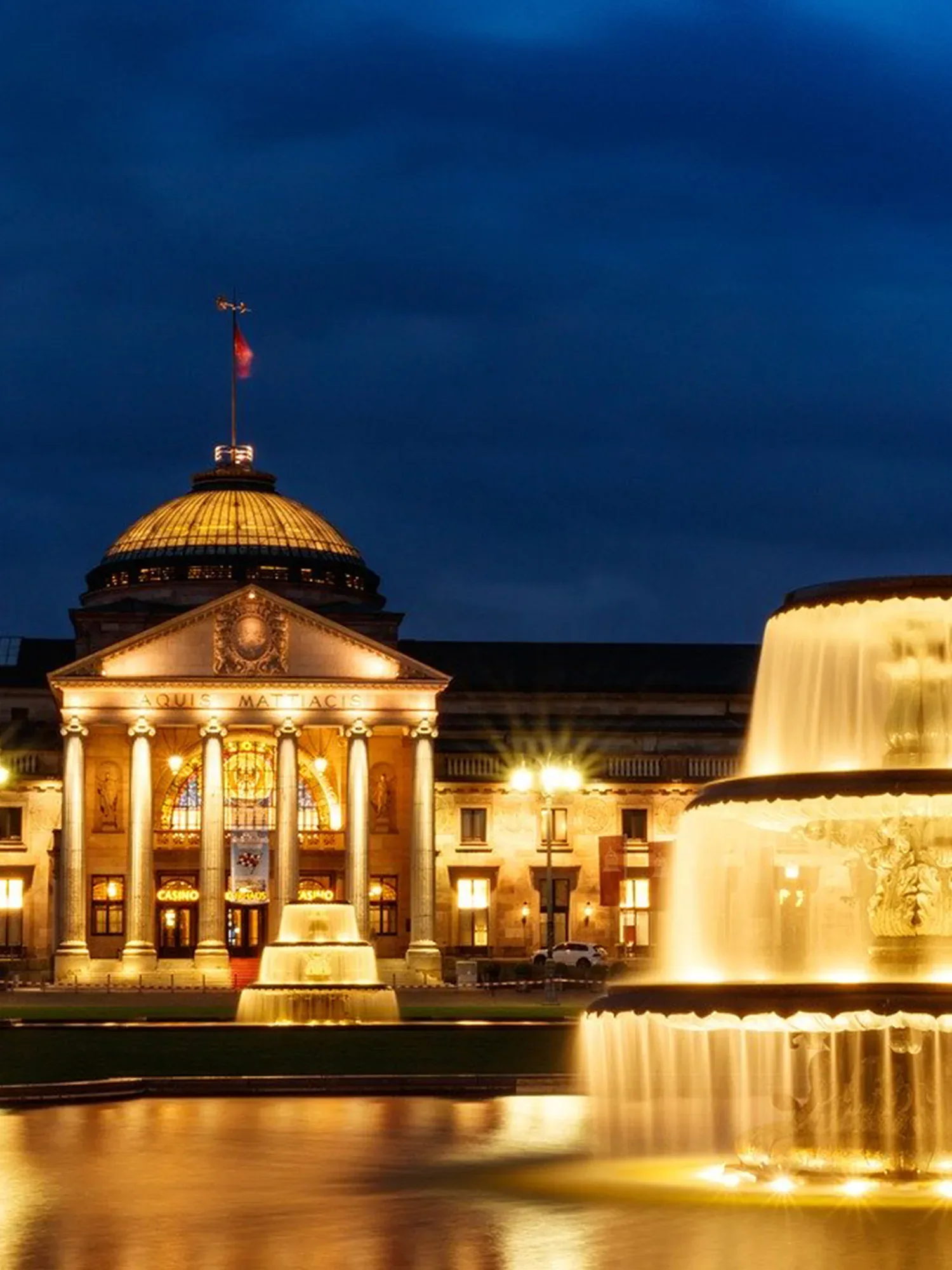 Nachtaufnahme des beleuchteten Kurhauses Wiesbaden mit majestätischer Fassade und beeindruckendem Springbrunnen im Vordergrund, der das Gebäude in warmem Licht reflektiert.