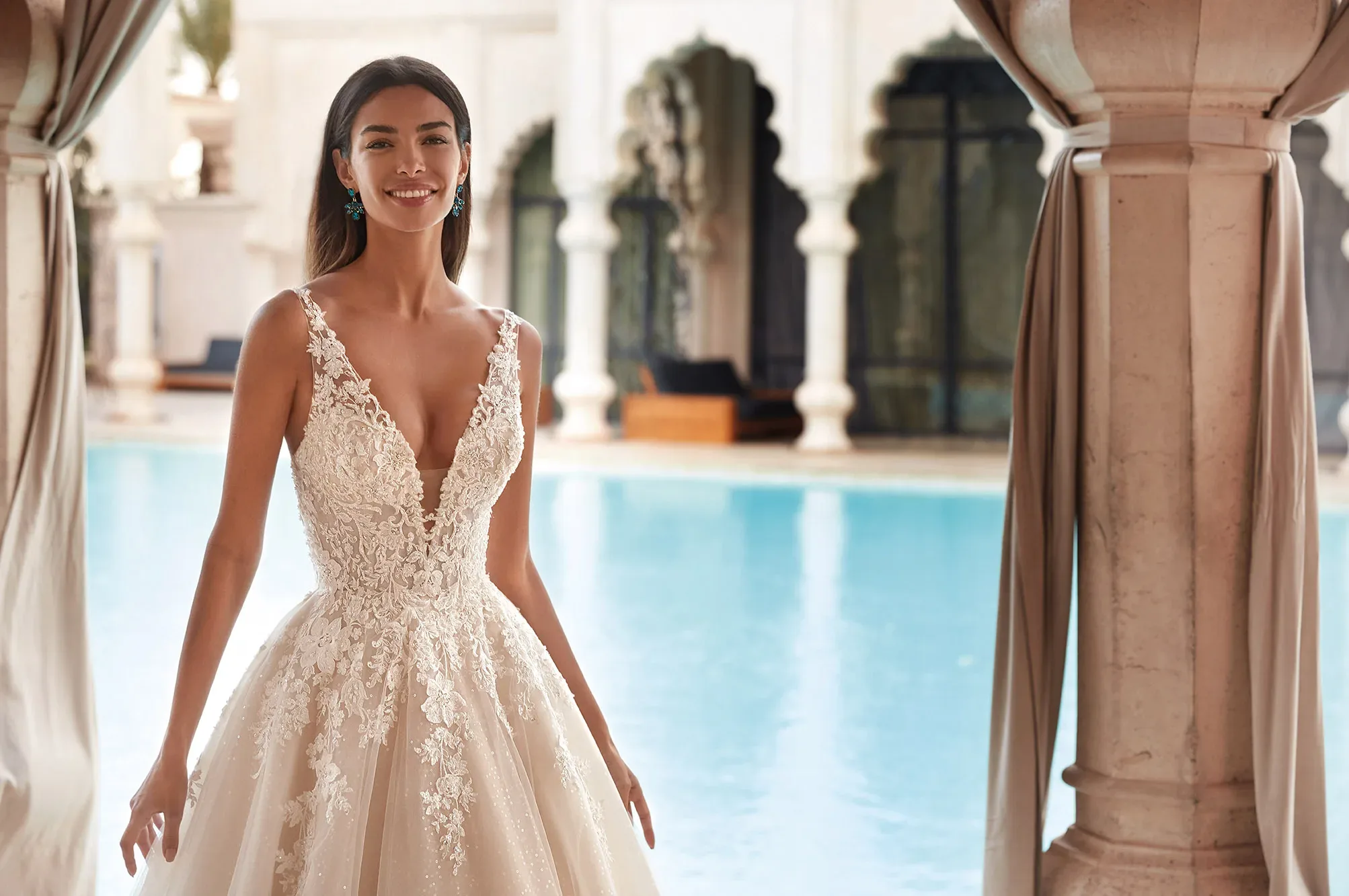 Braut in detailreichem Spitzenkleid mit tiefem Ausschnitt und floralen Applikationen, lächelnd vor einem orientalischen Pool.