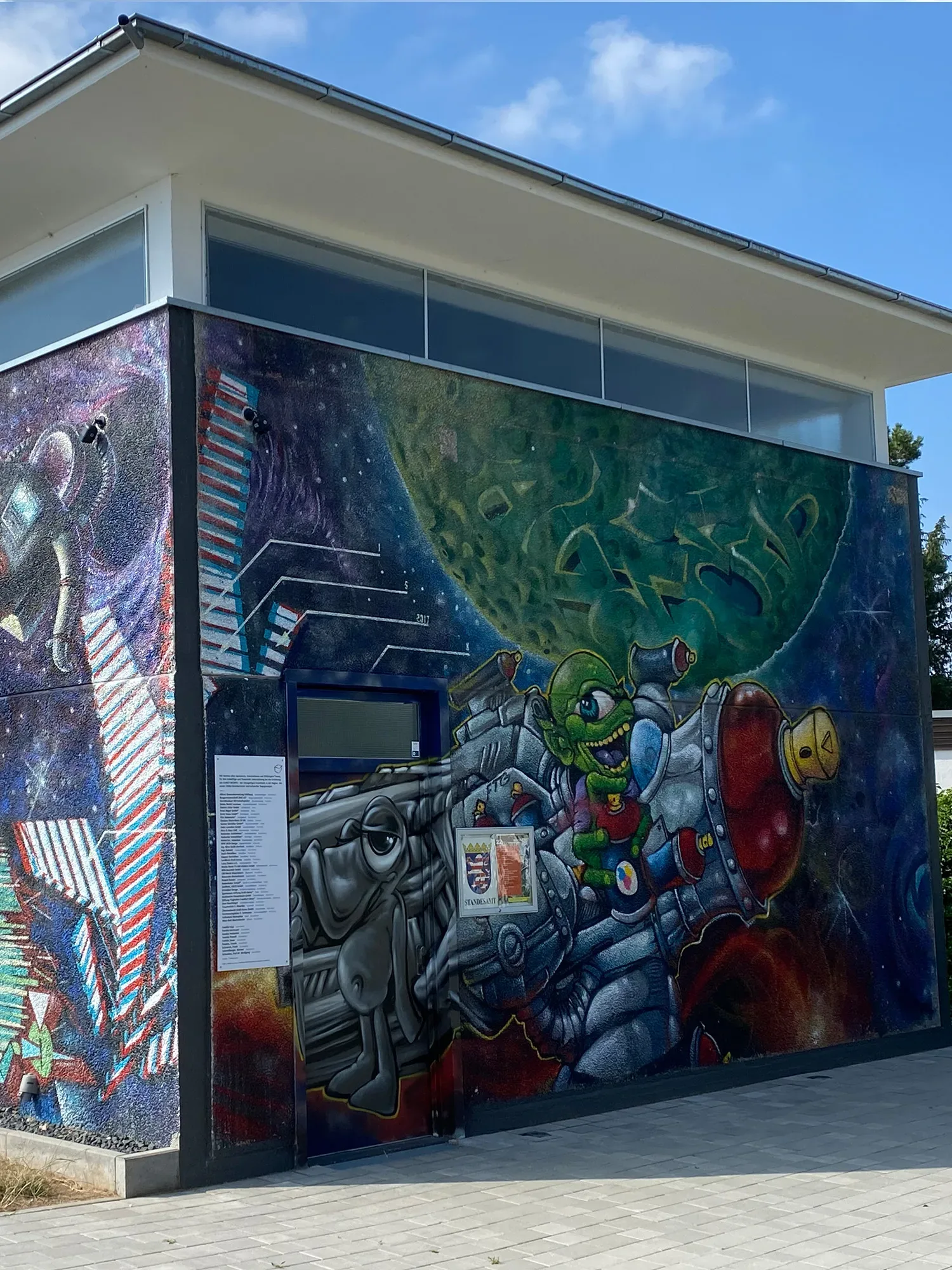 Außenwand des Bischofsheimer Kunstwürfels, dekoriert mit buntem Graffiti eines grünen Außerirdischen im Weltraum.