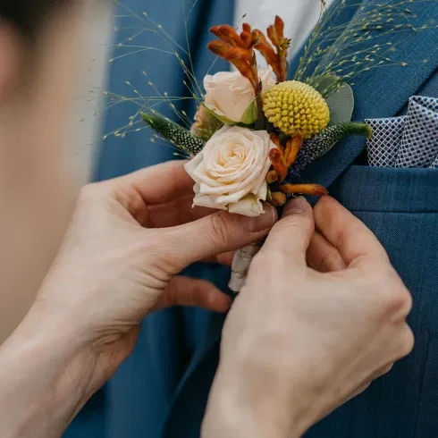 Eine Nahaufnahme zeigt Hände, die eine Boutonniere am Revers eines blauen Anzugs befestigen. Die Boutonniere besteht aus cremefarbenen Rosen, gelben Kugelblumen und orangenen Akzenten, ergänzt durch zarte Gräser.