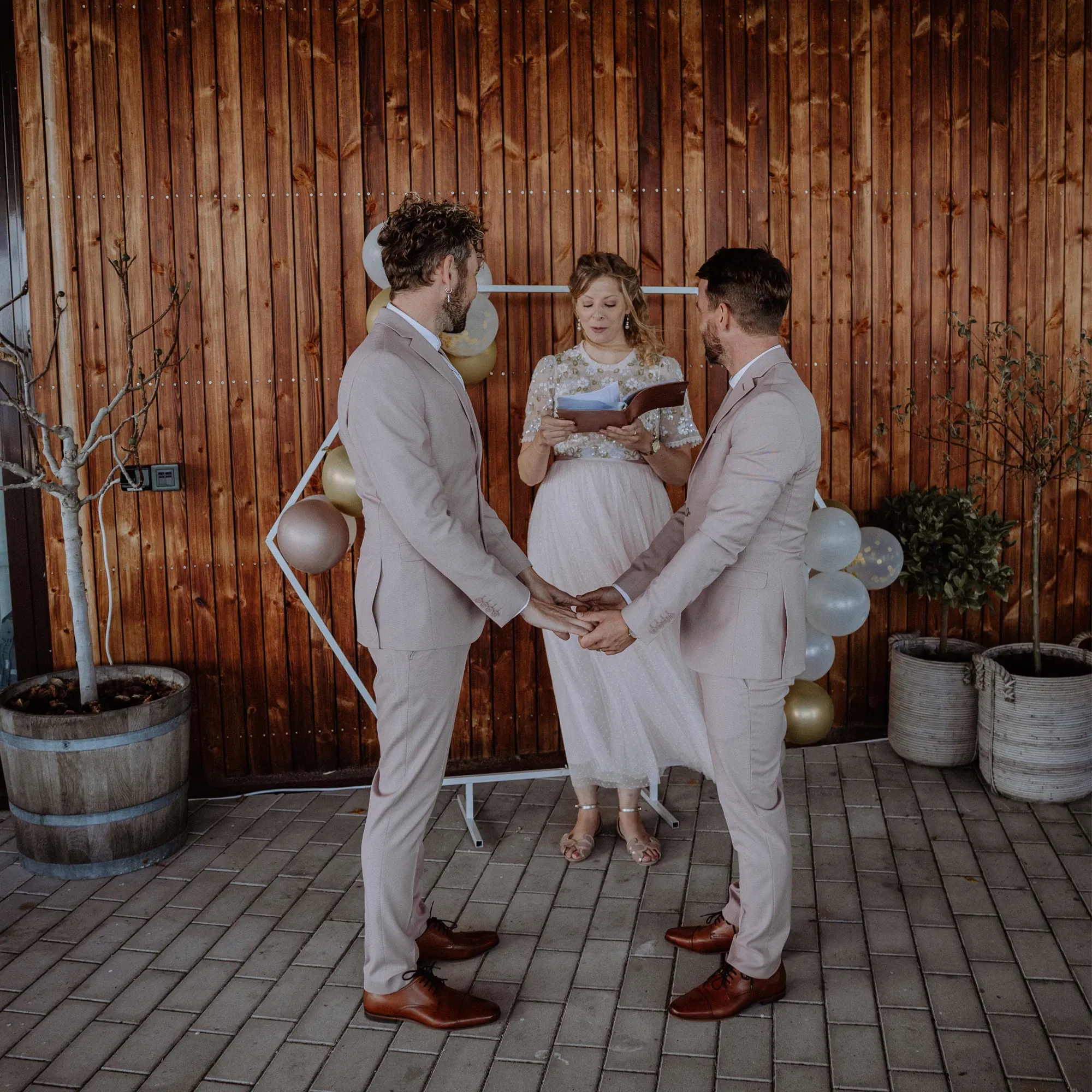 Ein gleichgeschlechtliches Paar in eleganten Anzügen hält sich während der Trauung die Hände, begleitet von einer Rednerin vor einer Holzwand.