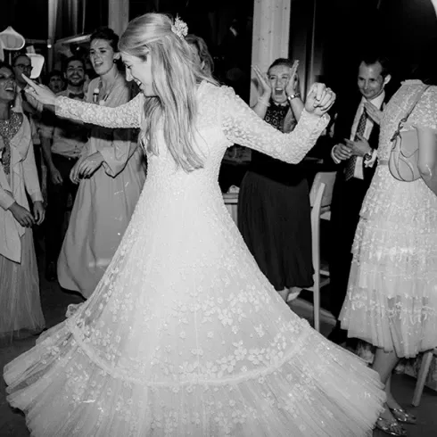 Braut tanzt glücklich in ihrem langen, bestickten Hochzeitskleid auf der Tanzfläche, umgeben von lächelnden Gästen, während sie sich elegant im Kreis dreht. Die Szene ist in schwarz-weiß gehalten, was die fröhliche und lebendige Atmosphäre der Hochzeit einfängt