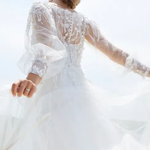 Rückansicht einer Braut in einem weißen Spitzenhochzeitskleid mit langen Ärmeln und einer aufwendigen Hochsteckfrisur, die im Wind steht, umgeben von sanftem Licht und einem klaren Himmel.