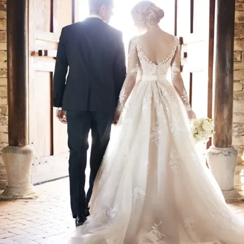 Ein Brautpaar schreitet Hand in Hand durch große Holztüren in einem rustikalen Gebäude. Die Braut trägt ein aufwendig besticktes weißes Kleid mit langer Schleppe, der Bräutigam einen dunklen Anzug.