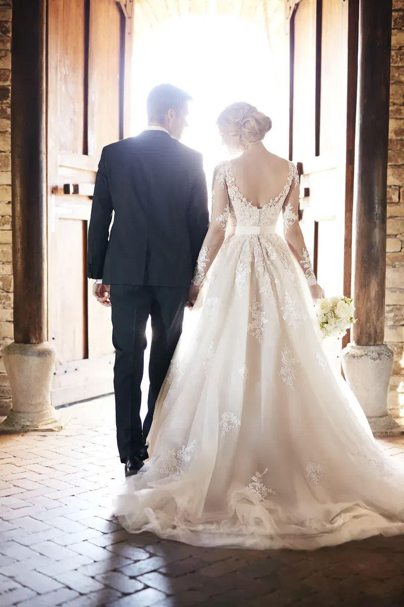 Ein Brautpaar schreitet Hand in Hand durch große Holztüren in einem rustikalen Gebäude. Die Braut trägt ein aufwendig besticktes weißes Kleid mit langer Schleppe, der Bräutigam einen dunklen Anzug.