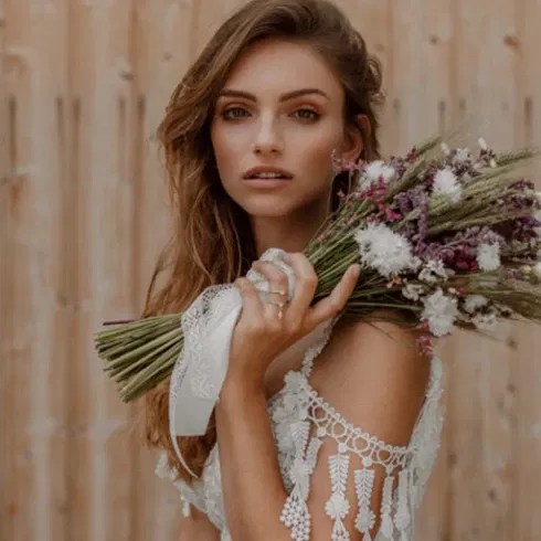 Eine Braut im Boho-Stil hält einen Strauß aus wilden, getrockneten Blumen und schaut mit einem intensiven Blick in die Kamera. Ihr langes Haar fällt sanft über ihre Schulter.