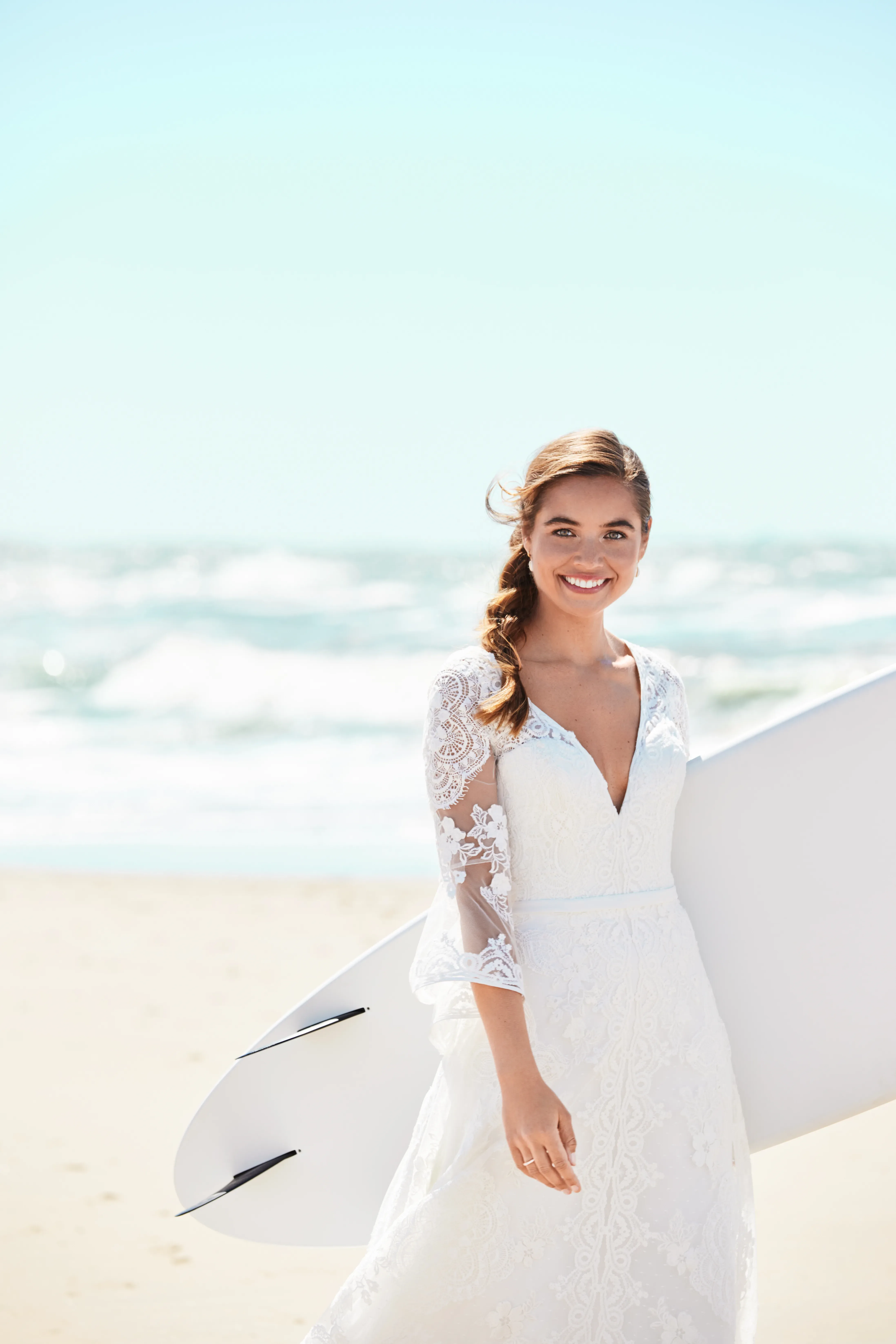 Eine strahlende Braut in einem weißen Spitzenkleid hält ein Surfbrett am Strand, bereit für eine unkonventionelle Hochzeitsfeier.