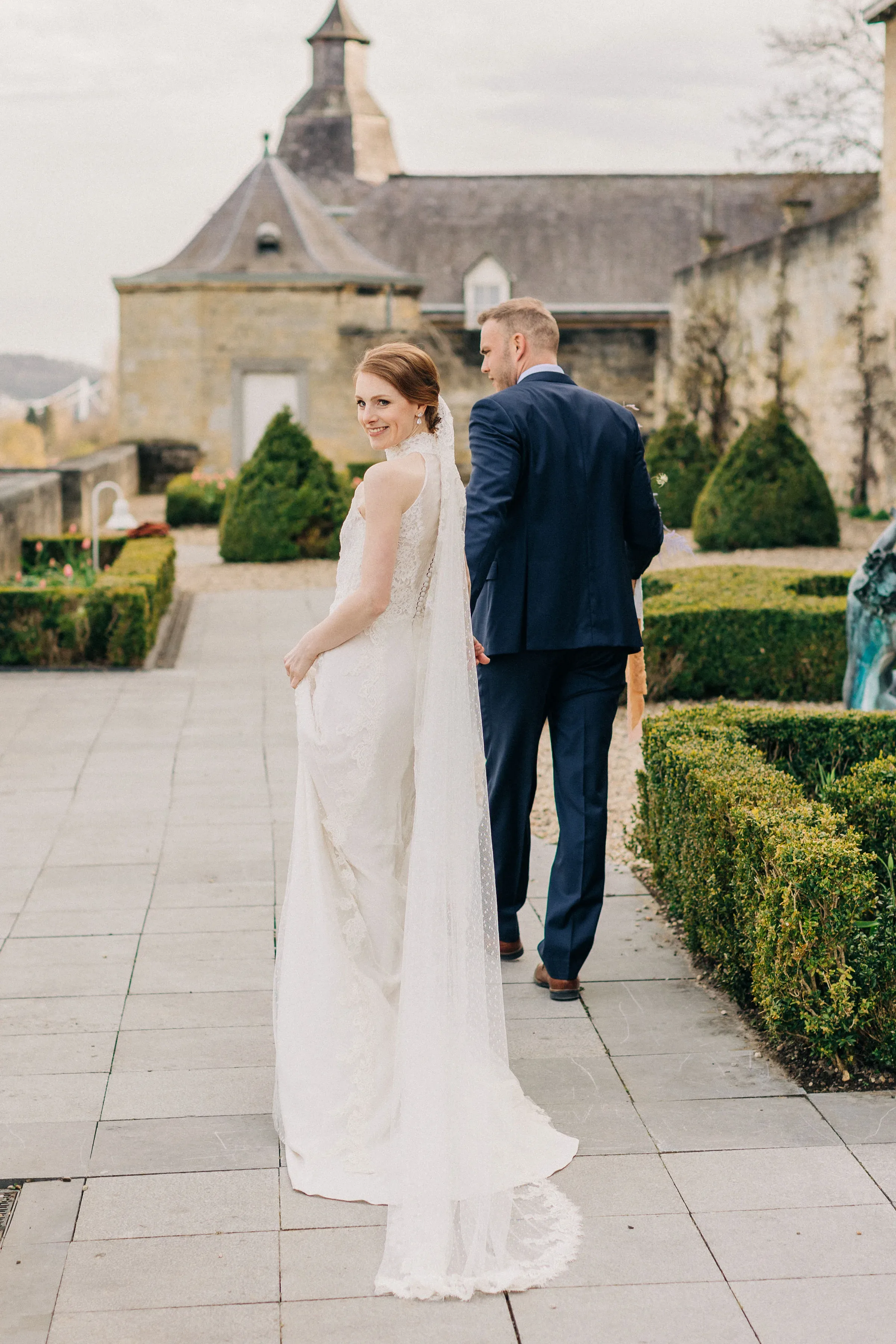 Ein Brautpaar spaziert durch einen formellen Garten vor einem historischen Gebäude. Die Braut trägt ein elegantes, spitzenbesetztes Kleid mit langem Schleier und schaut lächelnd über ihre Schulter zurück, während der Bräutigam in einem dunkelblauen Anzug an ihrer Seite geht.