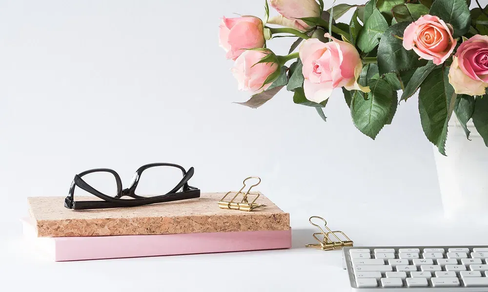 Ein stilvoll arrangierter Schreibtisch mit einem modernen weißen Hintergrund. Auf einem stapel aus zwei Büchern, eines mit einem Korkeinband und das andere rosa, liegt ein Paar schwarze Brillen. Daneben befinden sich zwei goldene Büroklammern. Am rechten Bildrand ist ein Teil einer Computertastatur zu sehen. Im Hintergrund sind rosa Rosen in einer weißen Vase, die eine frische und lebendige Atmosphäre schaffen.