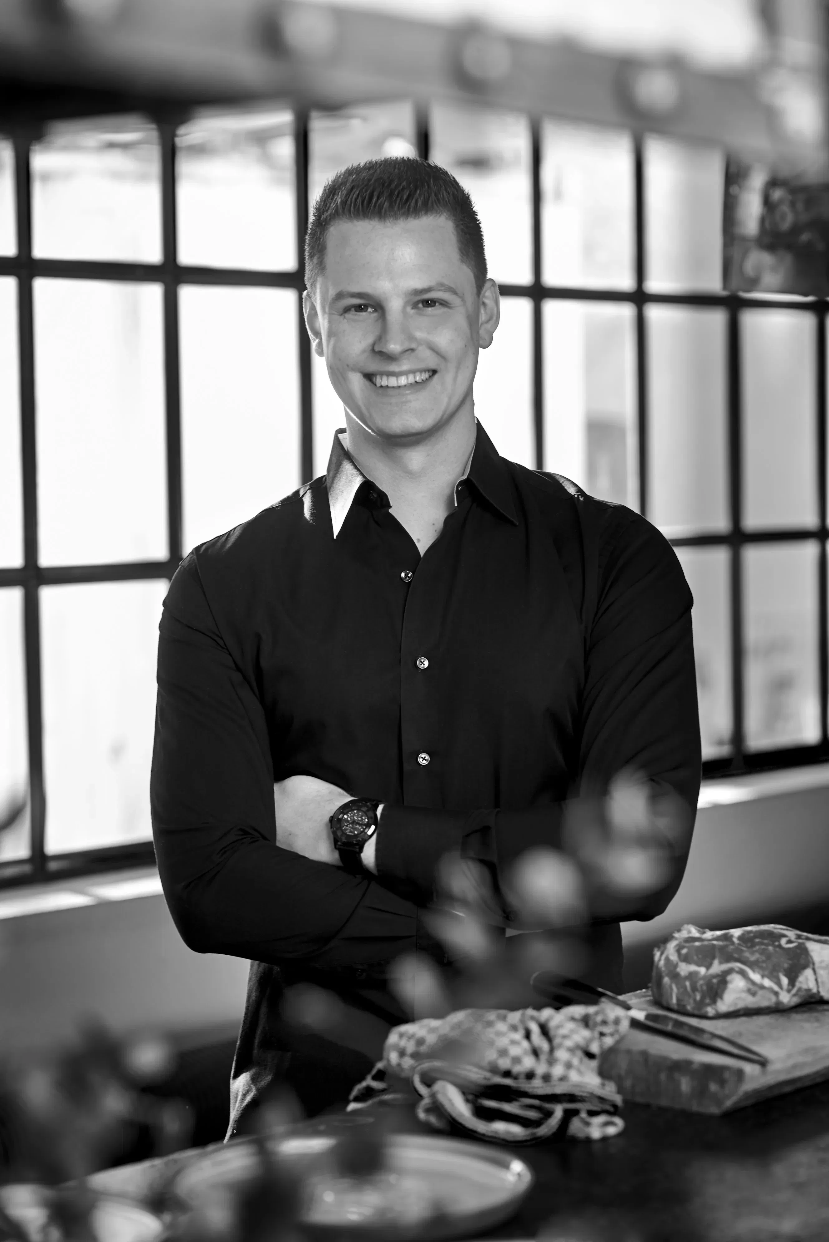 Schwarz-Weiß-Porträt eines Mannes in schwarzem Hemd, lächelnd mit verschränkten Armen vor einem Fenster stehend.