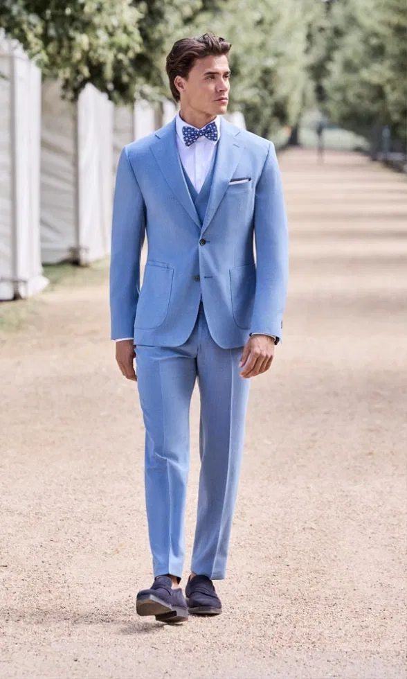 Mann in hellblauem Anzug mit passender Weste und gepunkteter Fliege geht einen Kiesweg entlang, umgeben von Bäumen und Zelten im Hintergrund.