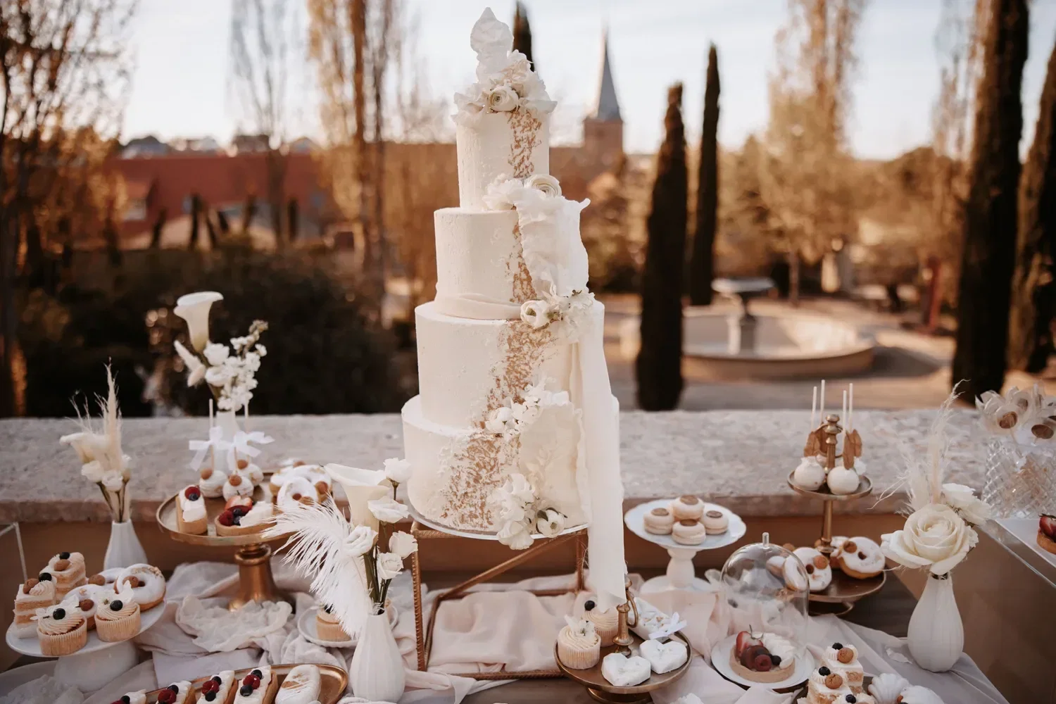Eleganter Desserttisch mit mehrstöckiger weißer Hochzeitstorte, umgeben von Blumen und süßen Leckereien im Freien dekoriert.