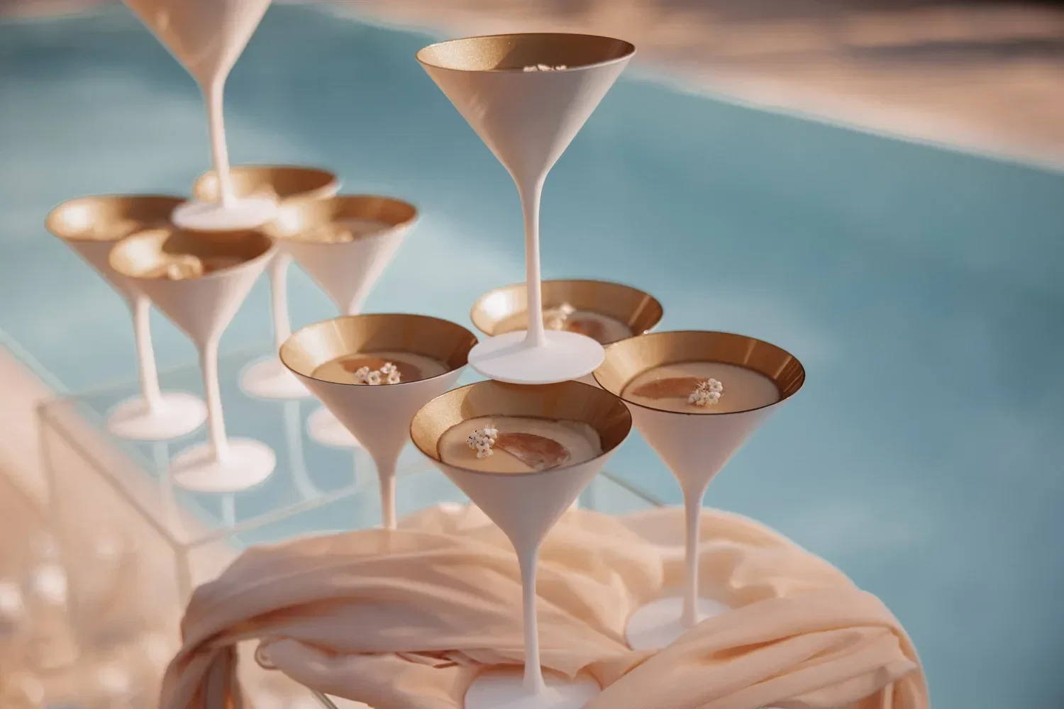 Ein Arrangement von stilvollen, matt-weißen Martini-Gläsern mit goldfarbenem Rand, dekorativ am Pool aufgestellt – ideal für eine elegante Sommerhochzeit.