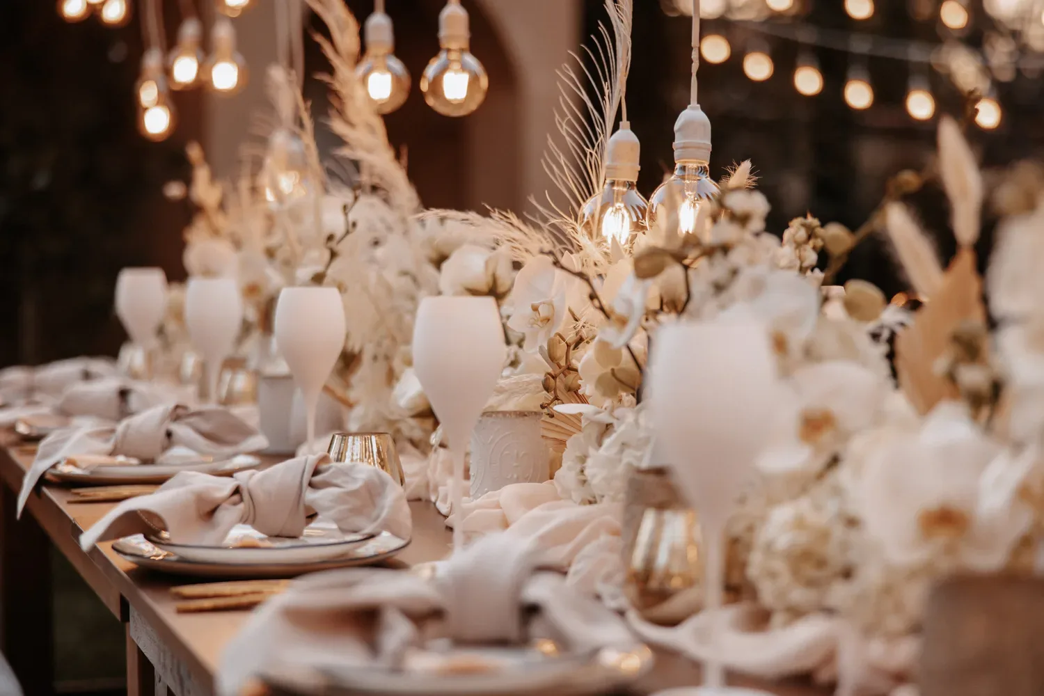 Festlich gedeckter Hochzeitstisch im Freien, dekoriert mit weißen Blumen, goldenen Akzenten und hängenden Glühbirnen für romantische Abendstimmung.