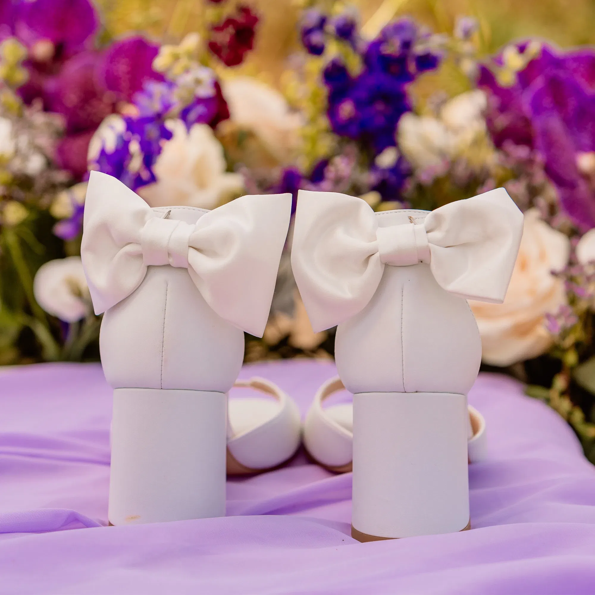 Das Bild zeigt die Rückseite eines Paares weißer Hochzeitsschuhe mit dicken Absätzen und großen Schleifen an der Ferse. Die Schuhe sind auf einem lila Tuch platziert, im Hintergrund sind Blumenarrangements aus Rosen und lila Blüten zu sehen, die eine romantische und festliche Atmosphäre erzeugen.