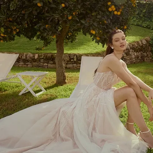 Braut in einem modernen, rosafarbenen Hochzeitskleid, sitzt entspannt auf einer Wiese unter einem Orangenbaum, umgeben von einem sommerlichen Gartenambiente.