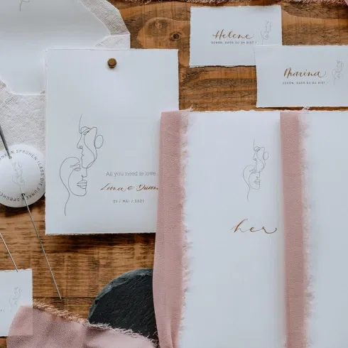 Stilvolle Hochzeitspapeterie mit feiner Kalligrafie und minimalistischen Illustrationen, dekoriert mit Stoffbändern auf einer rustikalen Holzoberfläche.