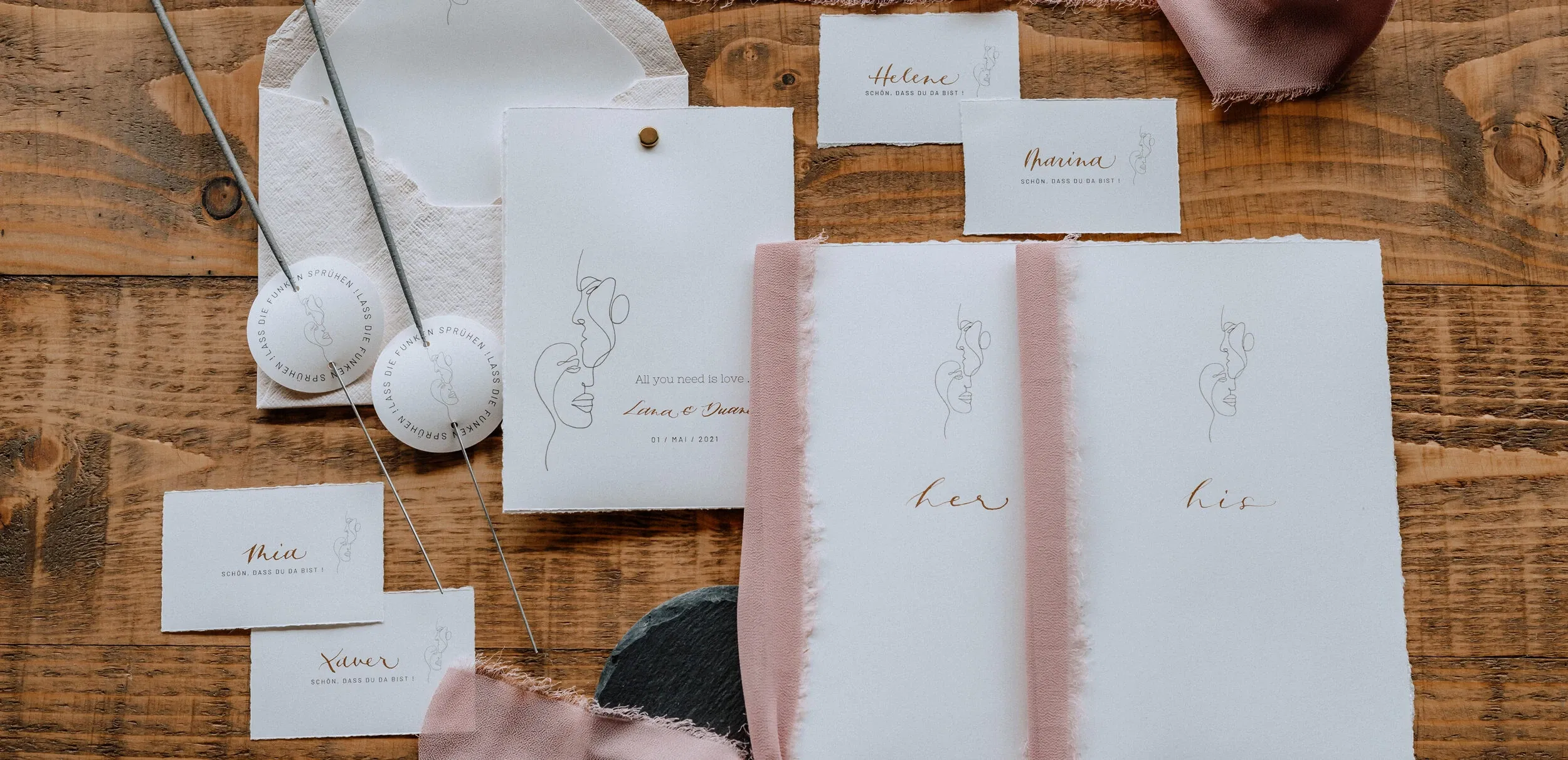Stilvolle Hochzeitspapeterie mit feiner Kalligrafie und minimalistischen Illustrationen, dekoriert mit Stoffbändern auf einer rustikalen Holzoberfläche.