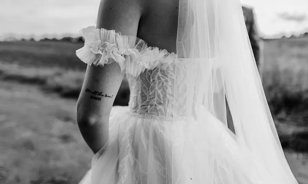 Eine Nahaufnahme der Rückseite einer Braut im Hochzeitskleid mit einem Tattoo am Arm, das den Text 'After all this time? ALWAYS' zeigt, aufgenommen im Freien.