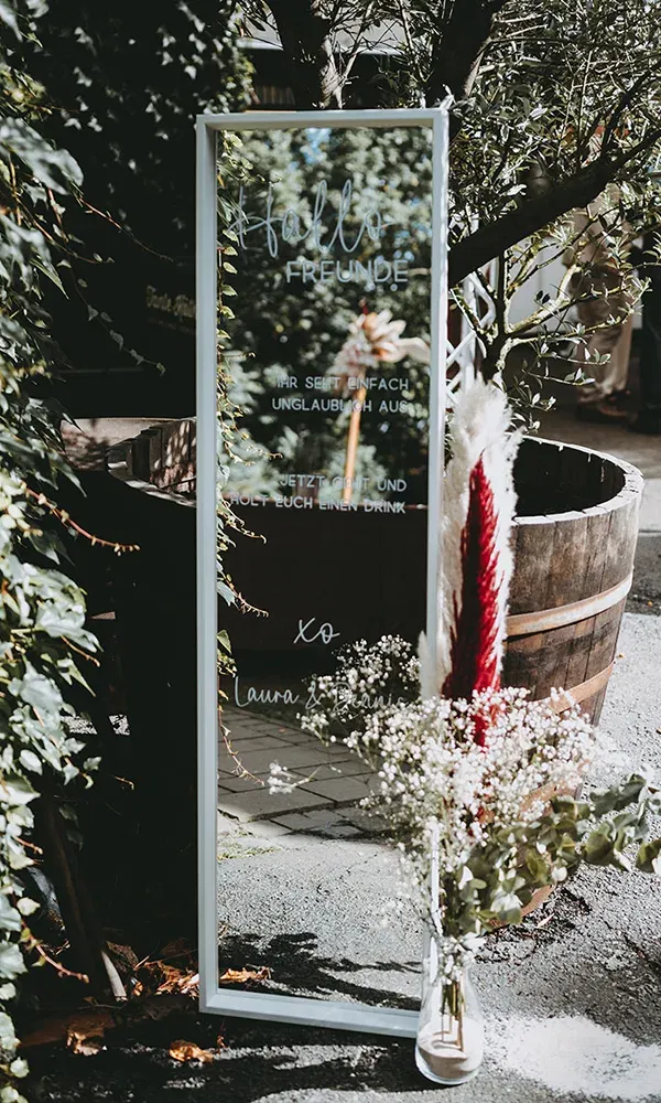 Ein dekorativer Spiegel auf einer Hochzeit, mit einer handgeschriebenen Nachricht: 'Hallo Freunde, ihr seht einfach unglaublich aus. Jetzt geht und holt euch einen Drink.' Verziert mit Blumen und Pampasgras.
