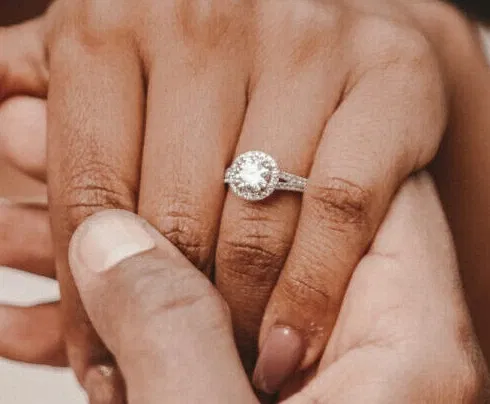Nahaufnahme eines funkelnden Verlobungsrings mit Halo-Fassung am Finger einer Frau, gehalten von der Hand ihres Partners.
