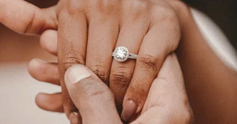 Nahaufnahme eines funkelnden Verlobungsrings mit Halo-Fassung am Finger einer Frau, gehalten von der Hand ihres Partners.