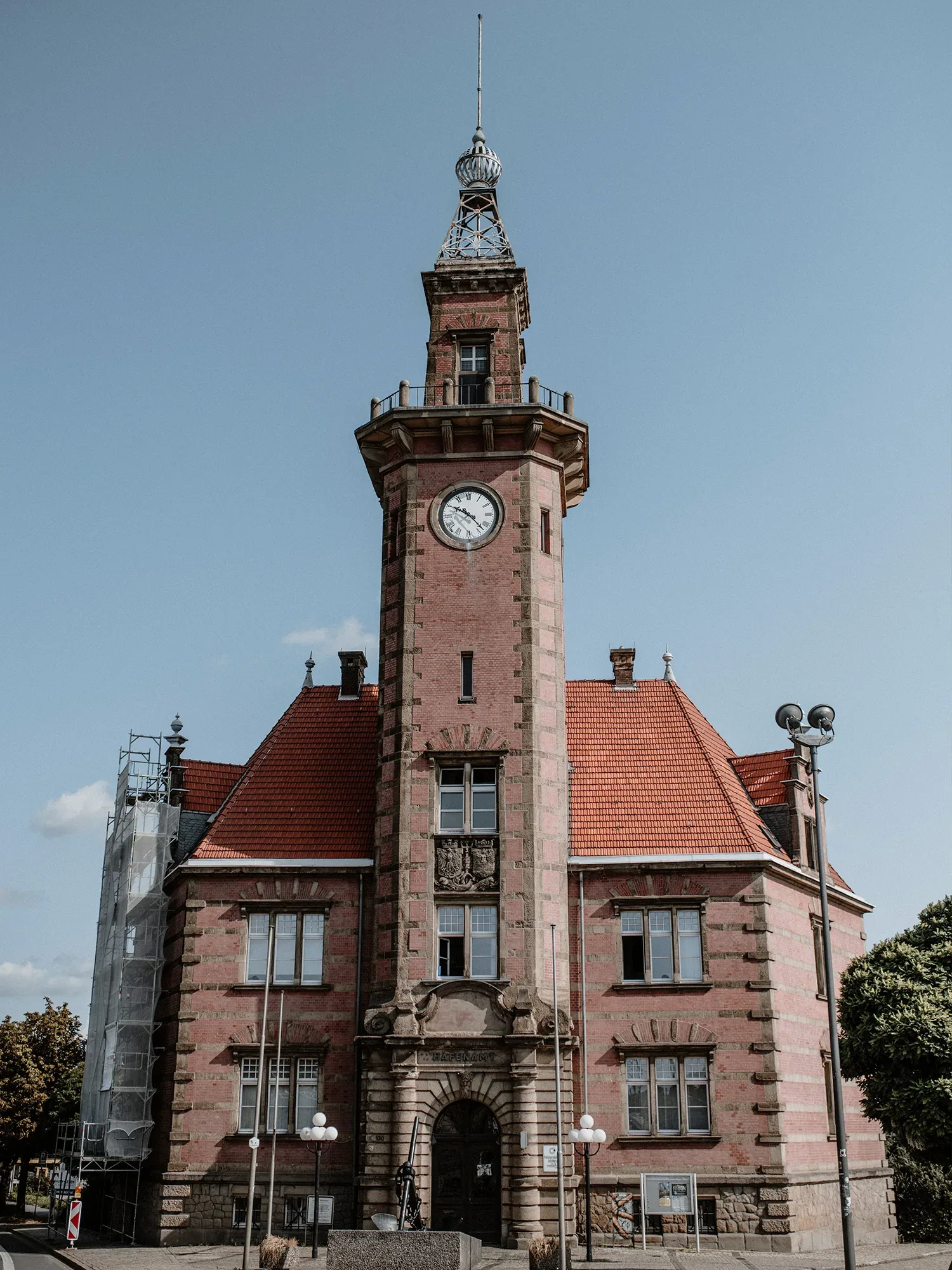 Backstein-Gebäude mit einem hohen Turm und Turmuhr, gekrönt von einer kunstvollen Metallspitze. Das Gebäude hat ein rotes Ziegeldach und verziertes Mauerwerk im Eingangsbereich. Die Fassade ist von Renovierungsgerüsten und Lampen umgeben.