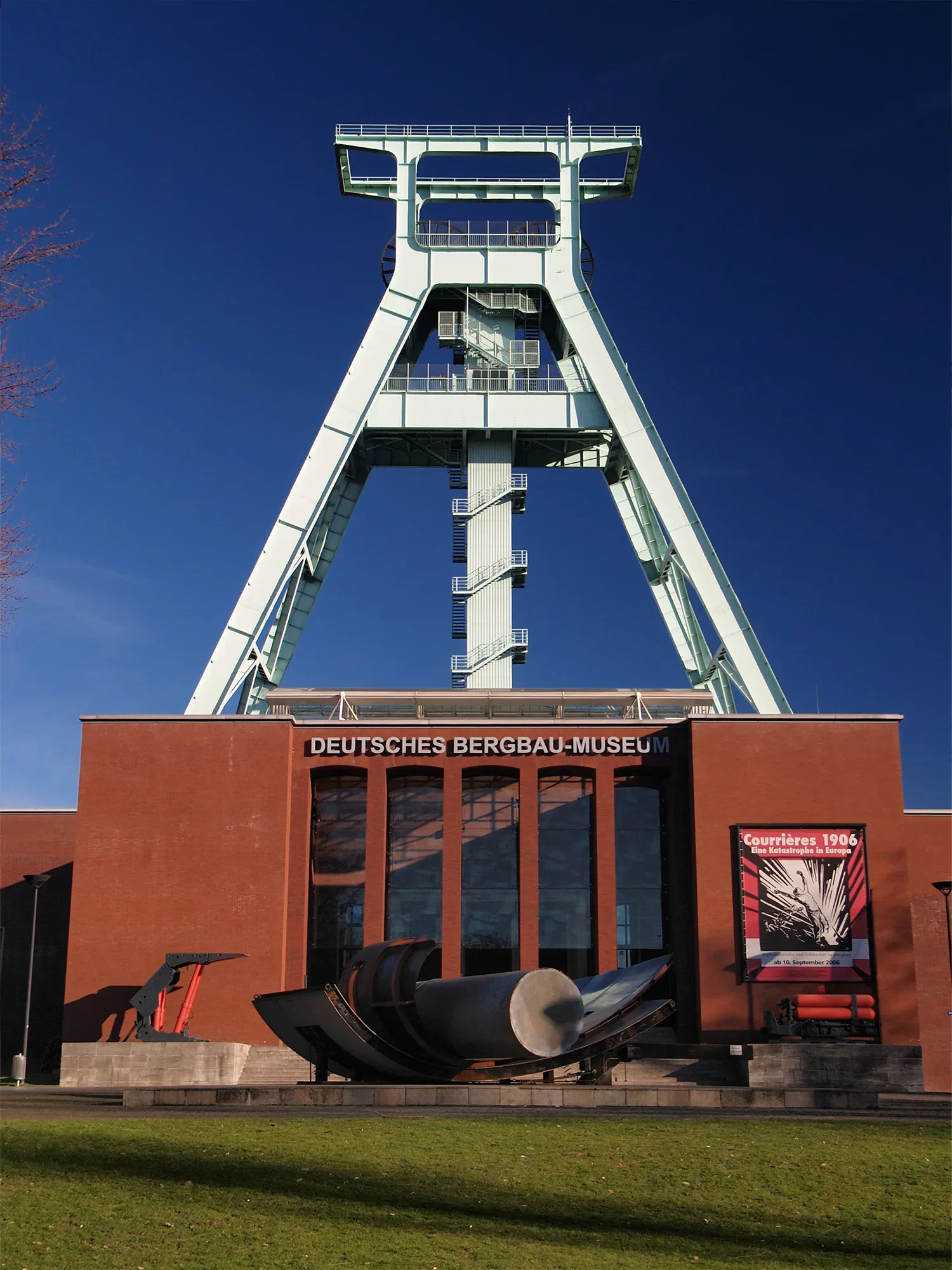 Das Deutsche Bergbau-Museum mit roter Backsteinfassade und grossem Förderturm vor strahlend blauem Himmel. Vor dem Gebäude befinden sich grosse Industriemaschinen, darunter eine Bergbauausrüstung aus Metall, und ein Werbeplakat für eine Ausstellung zum Thema „Courrières 1906“.
