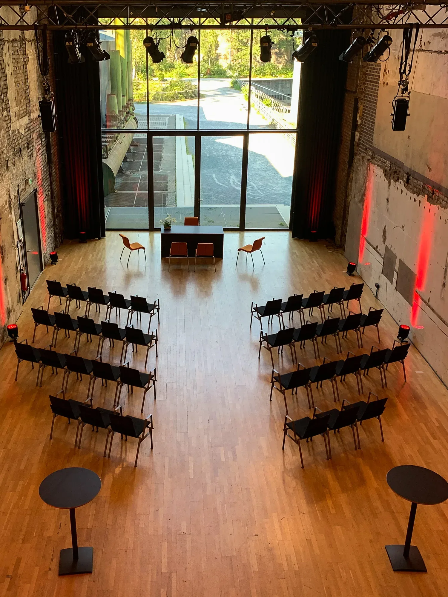 Ein moderner Tagungsraum mit grossen Fenstern, durch die Tageslicht auf die Holzböden fällt. Stuhlreihen sind in Reihen für eine Präsentation angeordnet, vorne steht ein Tisch mit zwei Stühlen. An den Wänden und Säulen sind rote Lichter installiert, die eine warme Atmosphäre schaffen.