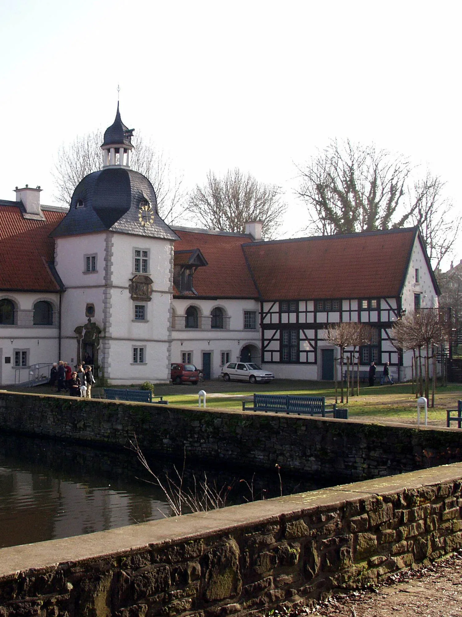 Ein historisches Gebäude mit weissem Turm, Uhr und rotem Ziegeldach, umgeben von einem Wassergraben. Im Vordergrund eine Steinmauer und Bänke in der gepflegten Gartenanlage. Ein Fachwerkbau schliesst rechts an das Hauptgebäude an.