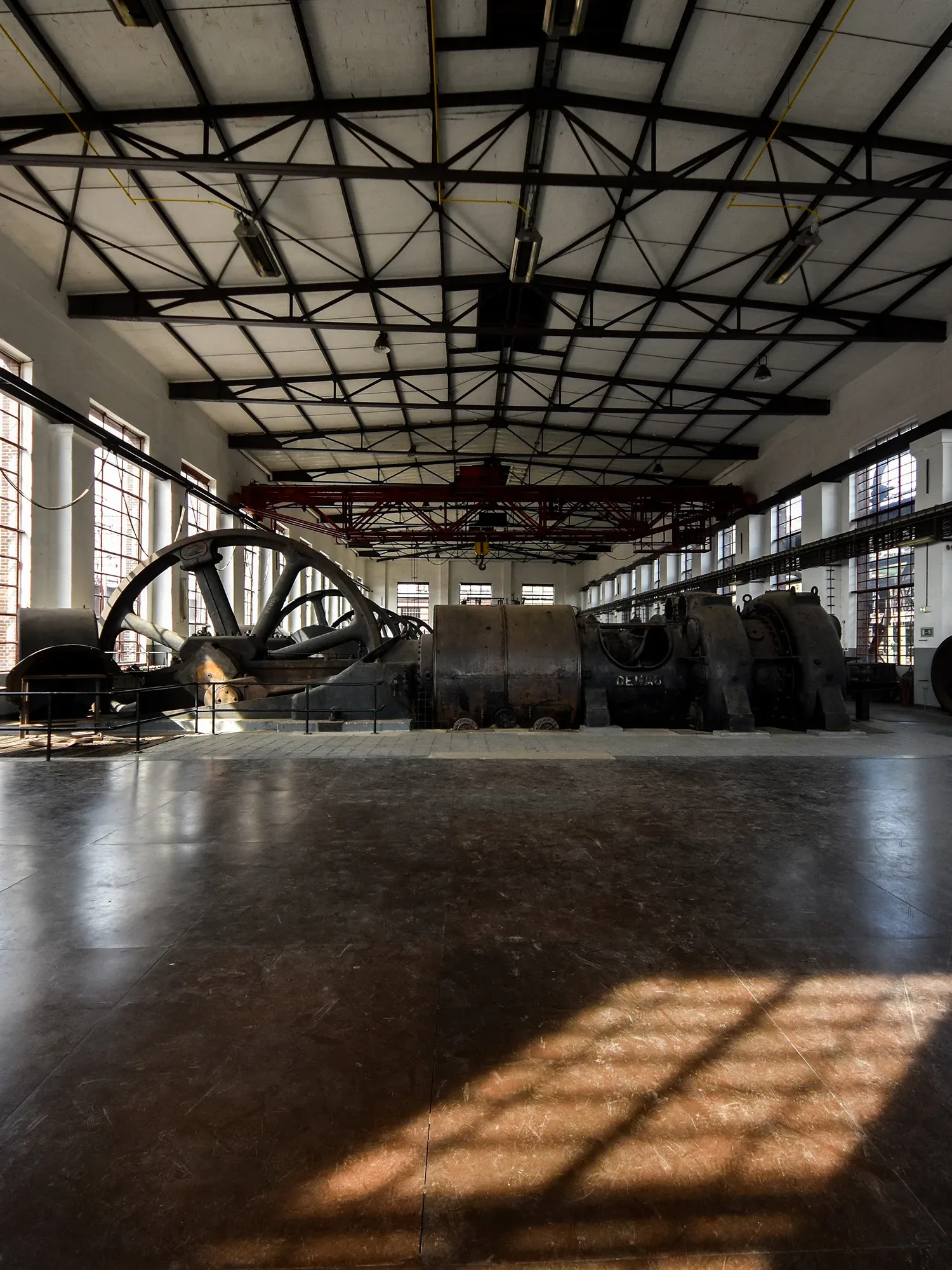 Weitläufige Industriehalle mit alten Maschinen und einem riesigen Schwungrad in der Mitte. Die hohen Fenster lassen Sonnenlicht herein, das Schatten auf den Boden wirft und die industrielle Atmosphäre verstärkt. Stahlträger an der Decke betonen die robuste Architektur.