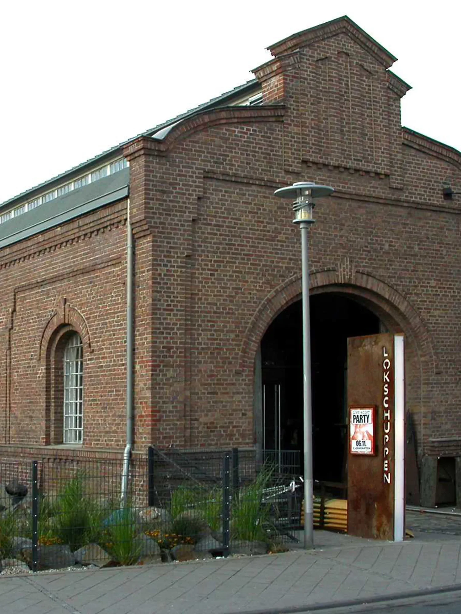 Historisches Gebäude aus Backstein mit der Aufschrift „Lokschuppen“ auf einem rostfarbenen Schild. Das Gebäude hat ein grosses Tor und Fenster im klassischen Industriestil. Im Vordergrund sind Gräser und kleine Steine als Landschaftsgestaltung zu sehen.