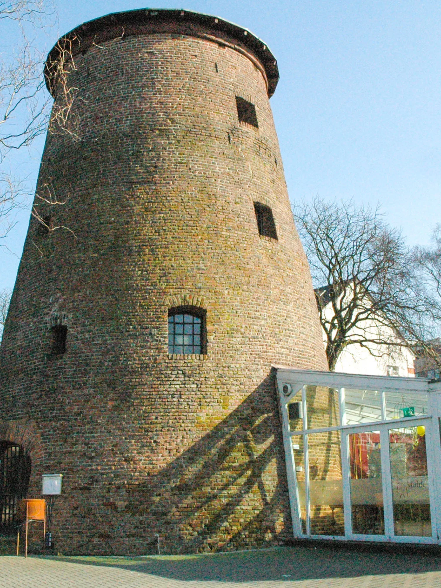 Historischer Backsteinturm mit kleinen Fenstern und abgerundetem Dach. Am Turm ist ein modernes, gläsernes Anbaugebäude angebracht. Die Schatten der Bäume fallen auf das Mauerwerk und betonen die Struktur der Steine.