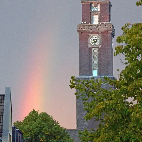 Hoher Kirchturm aus Backstein mit Uhr, vor einem regenverhangenen Himmel, durch den ein Regenbogen in sanften Farben leuchtet. Rechts des Turms und im Vordergrund sind grüne Bäume, die die Szene umrahmen.