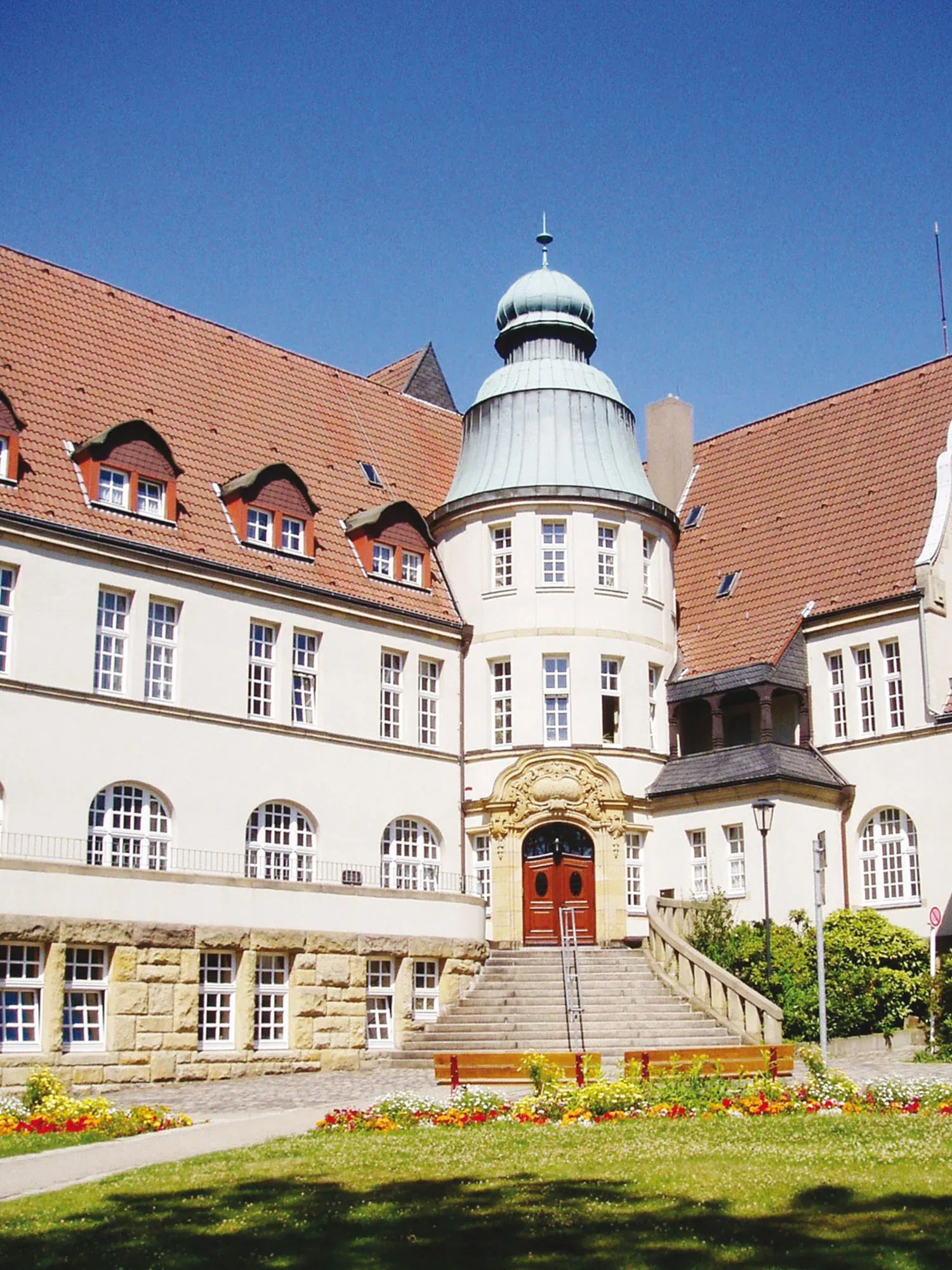Ein prächtiges historisches Gebäude mit rotem Ziegeldach und einer großen Kuppel über dem Eingang. Die weiße Fassade ist mit vielen Fenstern und einem steinernen Eingangsportal verziert. Im Vordergrund blüht ein farbenfrohes Blumenbeet im gepflegten Garten.